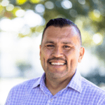 Close up of darkhaired man with mustache wearing a blue checkered shirt outside with a smile.